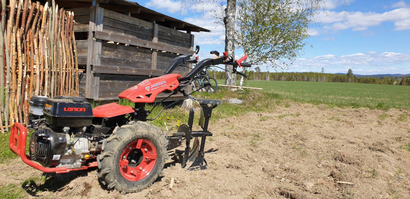 The red Jansen two-wheel tractor with the plough attached, standing on freshly worked soil next to a wooden shed